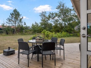 An outdoor deck with a round table and four chairs, surrounded by a wooden deck and a grassy area with trees.
