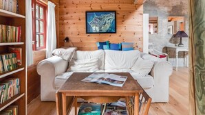 A cozy living room with a wooden ceiling, a white sofa, a coffee table with magazines, and a bookshelf filled with books.