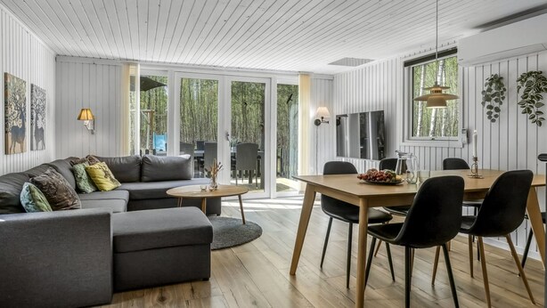 Una sala de estar moderna con sofá, mesa de comedor y sillas. Se ve una ventana grande con vista a árboles.