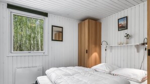 Un dormitorio con un ventanal grande, una cama con ropa de cama blanca, un armario de madera y una mesita de noche pequeña con una lámpara.