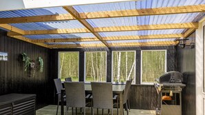 Un patio exterior con terraza de madera, mesa con sillas, una parrilla y ventanas amplias que ofrecen vistas a árboles.