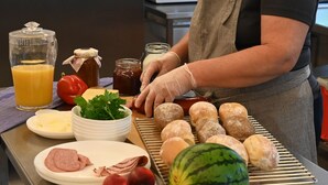 A kitchen with a person preparing food, including sausages, bread, and vegetables. - Sankt Anna Resort (Sankt Anna)
