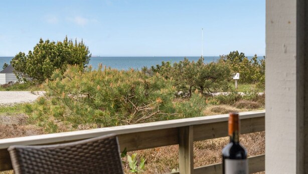 A wooden deck with a table and chairs, overlooking a beach with sparse vegetation and a clear view of the ocean.