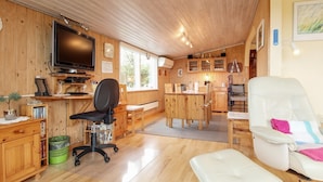 Ein gemütliches Blockhaushaus-Interieur mit Holzvertäfelung, einem Fernseher, einem Essbereich und einem weißen Stuhl mit farbenfroher Sitzkissen.