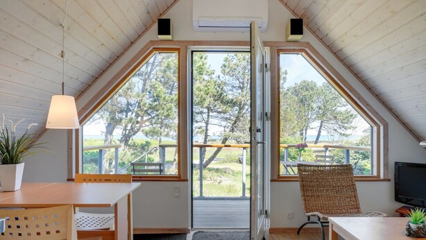 A cozy, well-lit room with a sloped ceiling, large windows offering a view of the outdoors, a dining area with a table and chairs, and a television on the right.