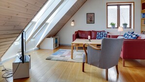 A cozy loft-style living room with a sloped ceiling, wooden beams, a red leather sofa, a grey armchair, a wooden coffee table, and a window with potted plants.