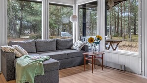 A cozy room with a grey sofa, a small wooden table, and a view of a forest through large windows.