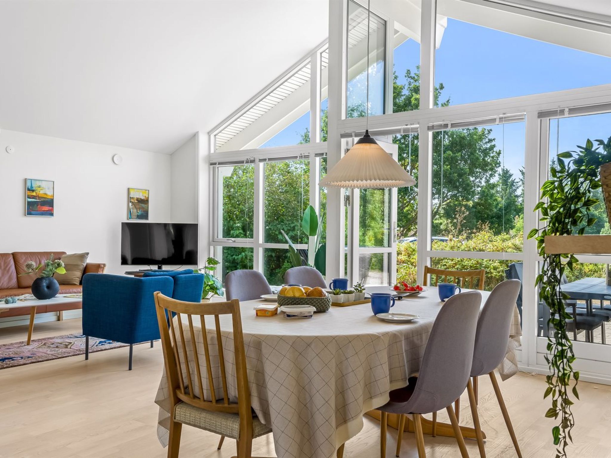 A modern living room with a dining area, large windows, and a view of greenery.