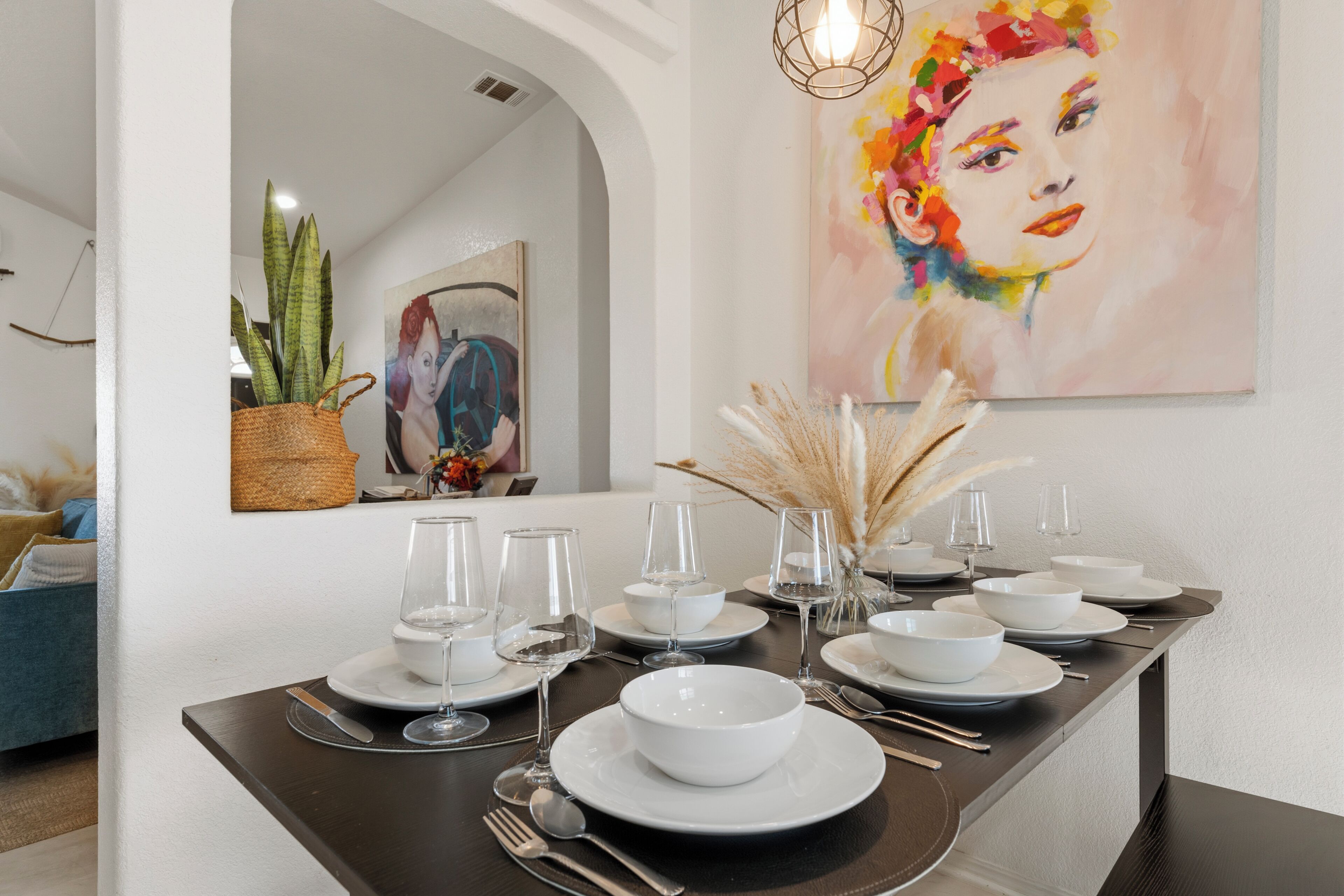A dining table set with white plates, bowls, and glasses, a vase with dried plants, and a painting on the wall.