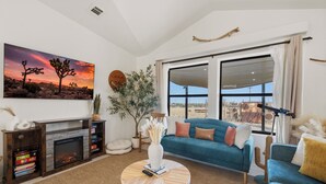 A living room with a fireplace, a large window, a blue sofa, and a desert landscape painting. - Summer Relax & Unwind @ Genesis Oasis - Wifi + Spa! Pet Friendly! (Joshua Tree)