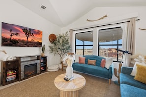 A living room with a fireplace, a large window, a blue sofa, and a desert landscape painting.
