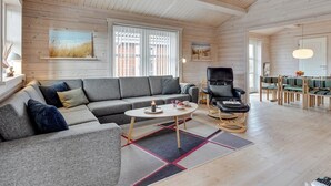 Amplia sala de estar con un sofá modular gris, una mesa de centro redonda y un sillón de cuero negro. Incluye una zona de comedor con mesa y sillas, y un ventanal con persianas.