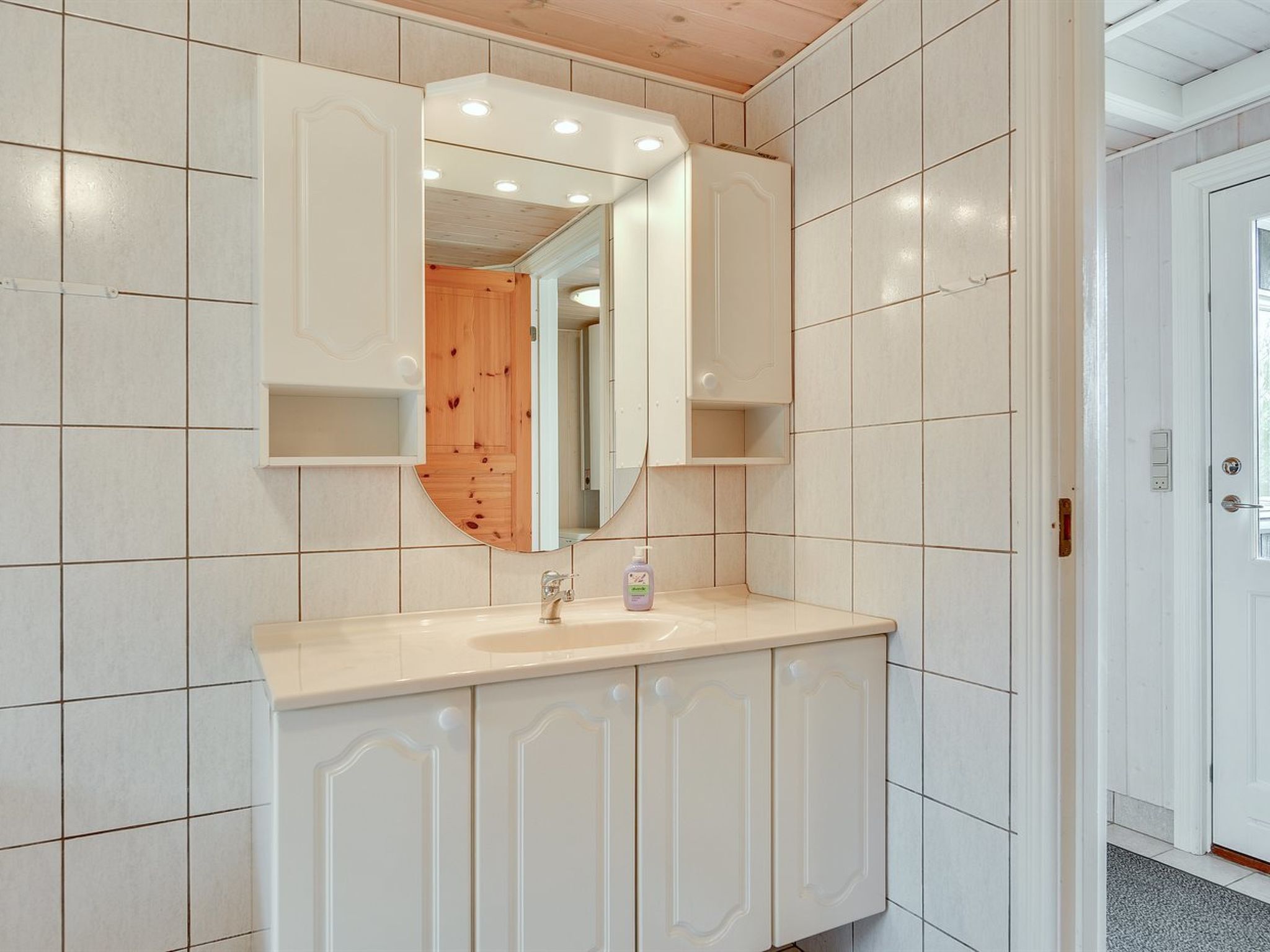 Ein Badezimmer mit weiß gefliesten Wänden, einer Holztür, einem Spiegel, einem Waschbecken und einem Schrank.
