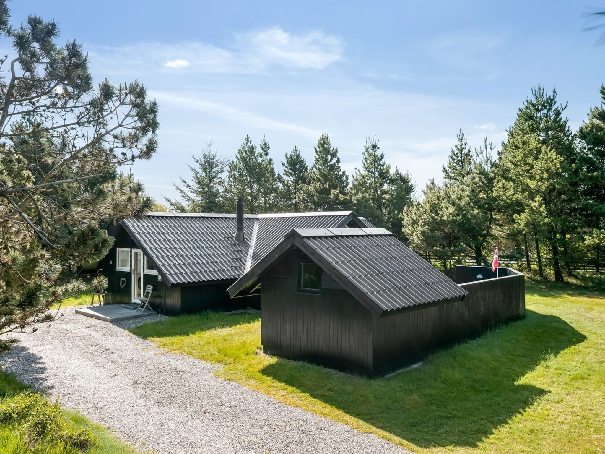 A modern cabin with a sloped roof, surrounded by a gravel driveway and lush greenery.