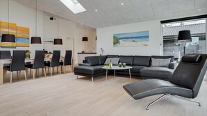 Un salon moderne avec un canapé gris, un coin repas avec des chaises, une table basse et un tableau représentant une plage.