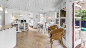 Um quarto amplo com piso de madeira, paredes brancas e uma janela grande. Há um espaço de refeições com mesa e cadeiras, e uma cozinha com bancada e armários.