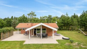 Une maison de plain-pied en briques, avec un toit en tuiles, une terrasse couverte comprenant une table et des chaises, et un portique à balançoire dans le jardin.