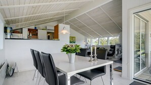 Un comedor moderno con una mesa blanca y sillas negras, un jarrón con flores y una cocina al fondo.