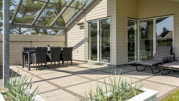 Un patio exterior moderno con cerramiento de vidrio, una zona de comedor con muebles de mimbre y una sala de estar con tumbonas.