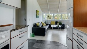 Una cocina moderna con armarios blancos, encimera negra y horno empotrado. Hay un sofá de cuero negro con cojines y una pequeña mesa redonda con un florero. Amplios ventanales ofrecen luz natural y vistas al exterior con vegetación.