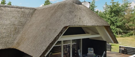 A modern house with a thatched roof, a deck with outdoor furniture, and a large glass door leading to an interior space.