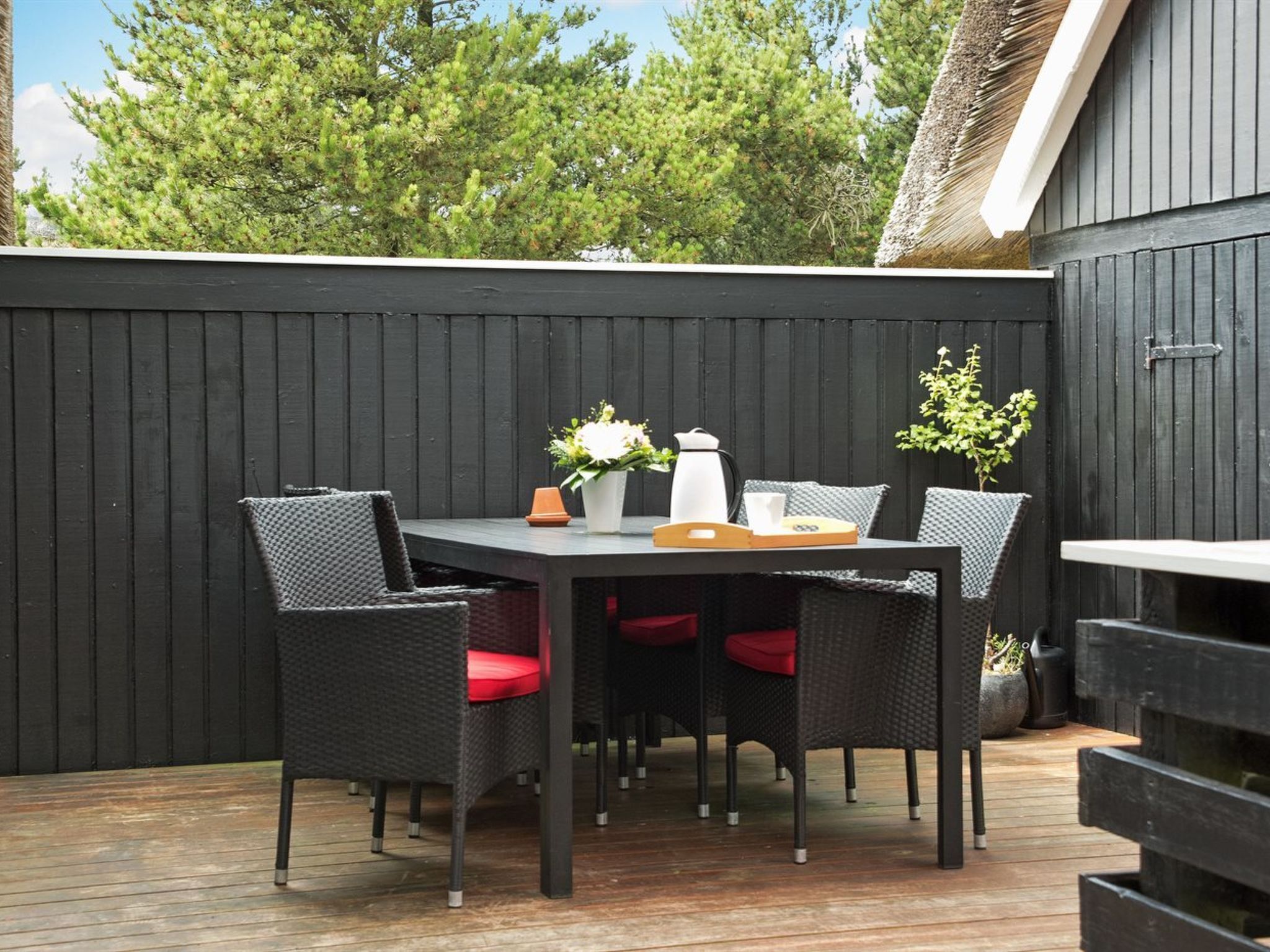 A patio with a dark wooden table and chairs, a white vase with flowers, and a coffee pot.