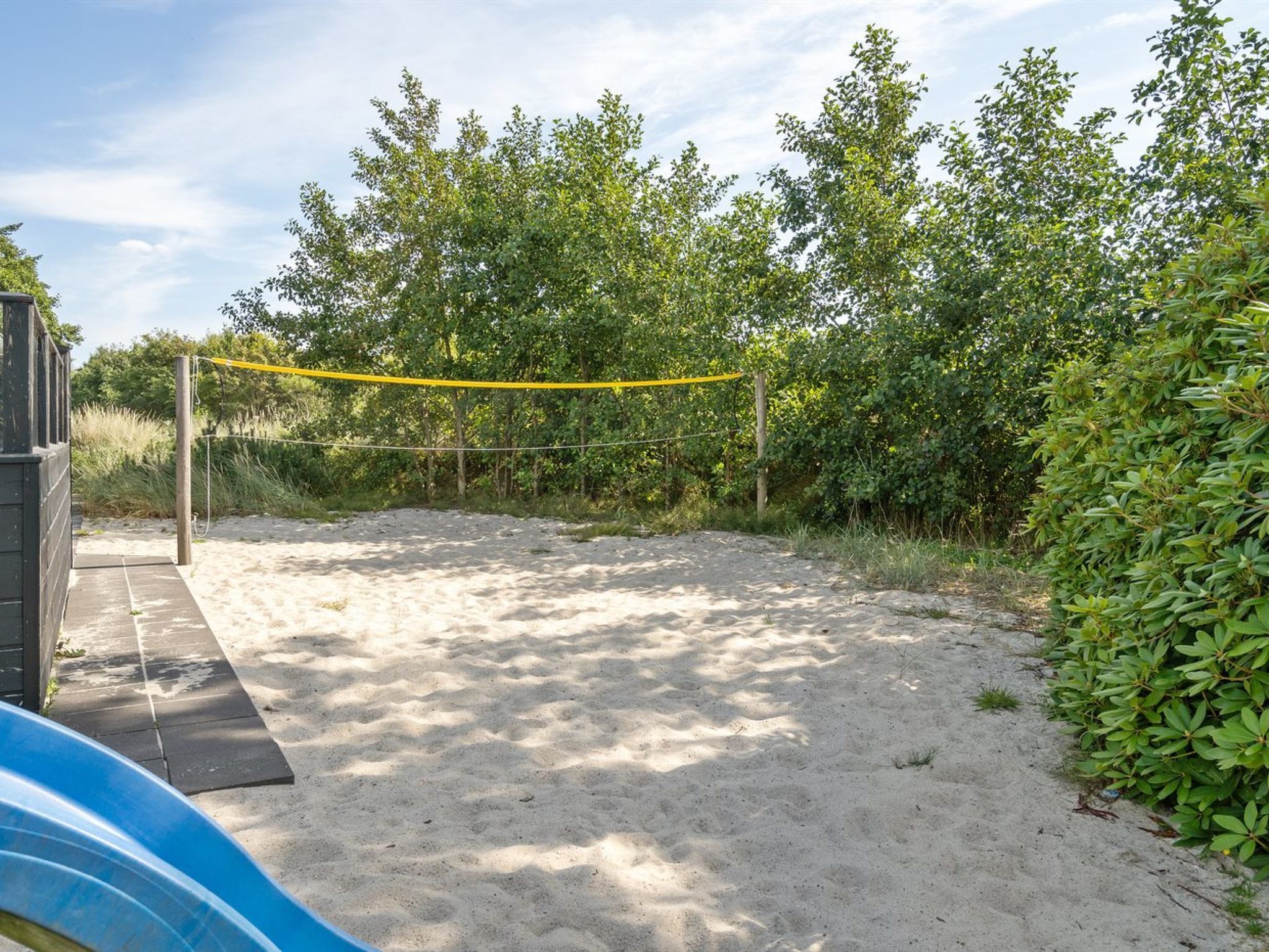Un'area sabbiosa all'aperto con una rete da pallavolo, circondata da una vegetazione lussureggiante e una struttura in legno.