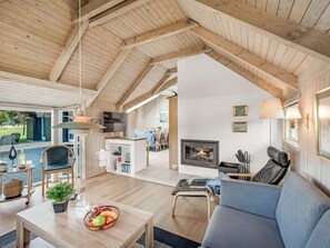 Uma sala de estar ampla com teto alto, vigas de madeira, lareira, área de jantar e vista para o exterior.