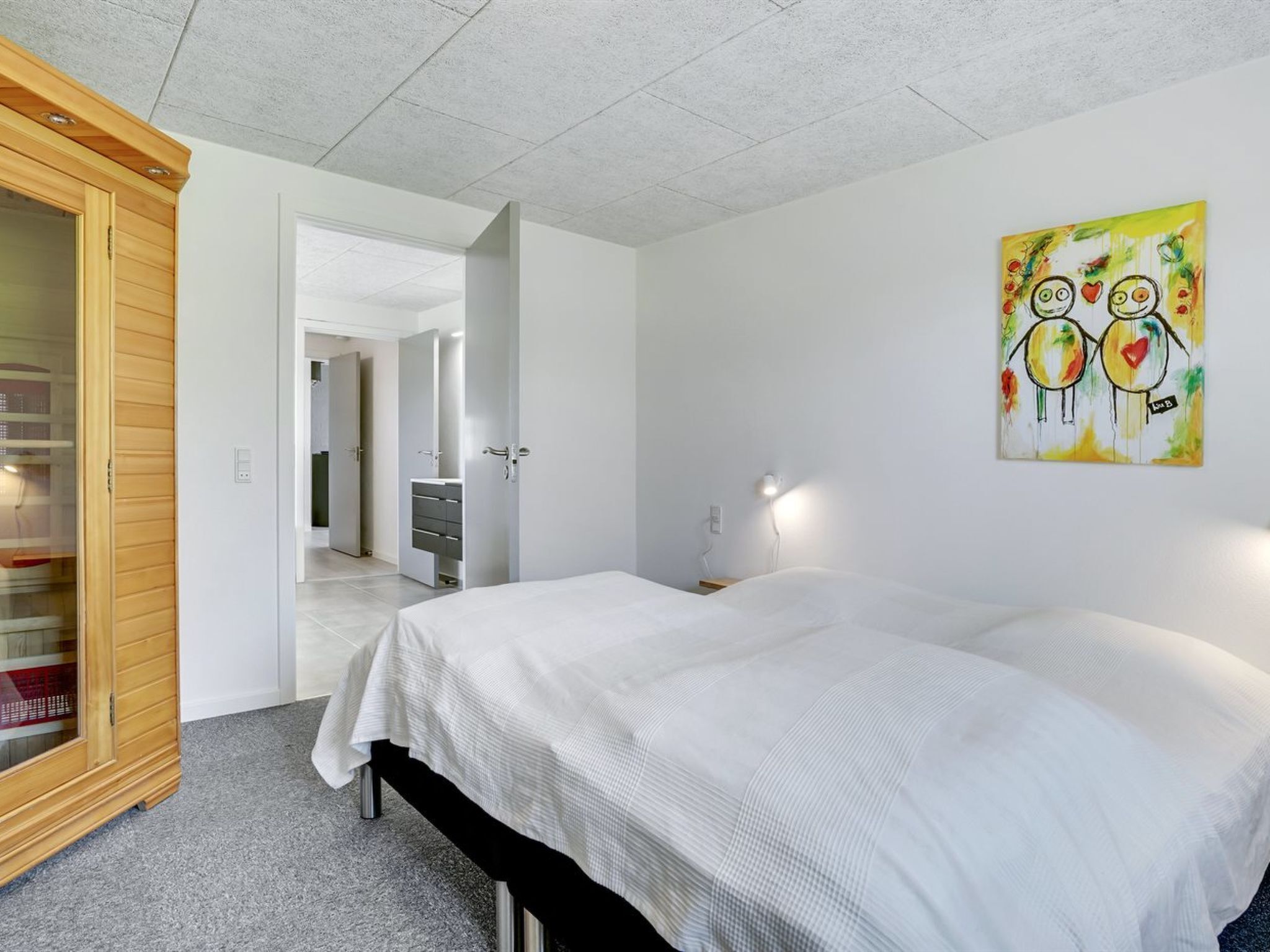 A bedroom with two beds, a wooden wardrobe, a bathroom with a mirror, and a colorful painting on the wall.