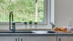 Cuina amb un aixeta d'acer inoxidable, una finestra que dona a vistes de vegetació, i un taulell amb plantes en test i pa.