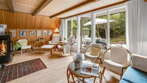 A cozy room with wooden walls and ceiling, a fireplace, comfortable seating, and large windows offering a view of the outdoors.