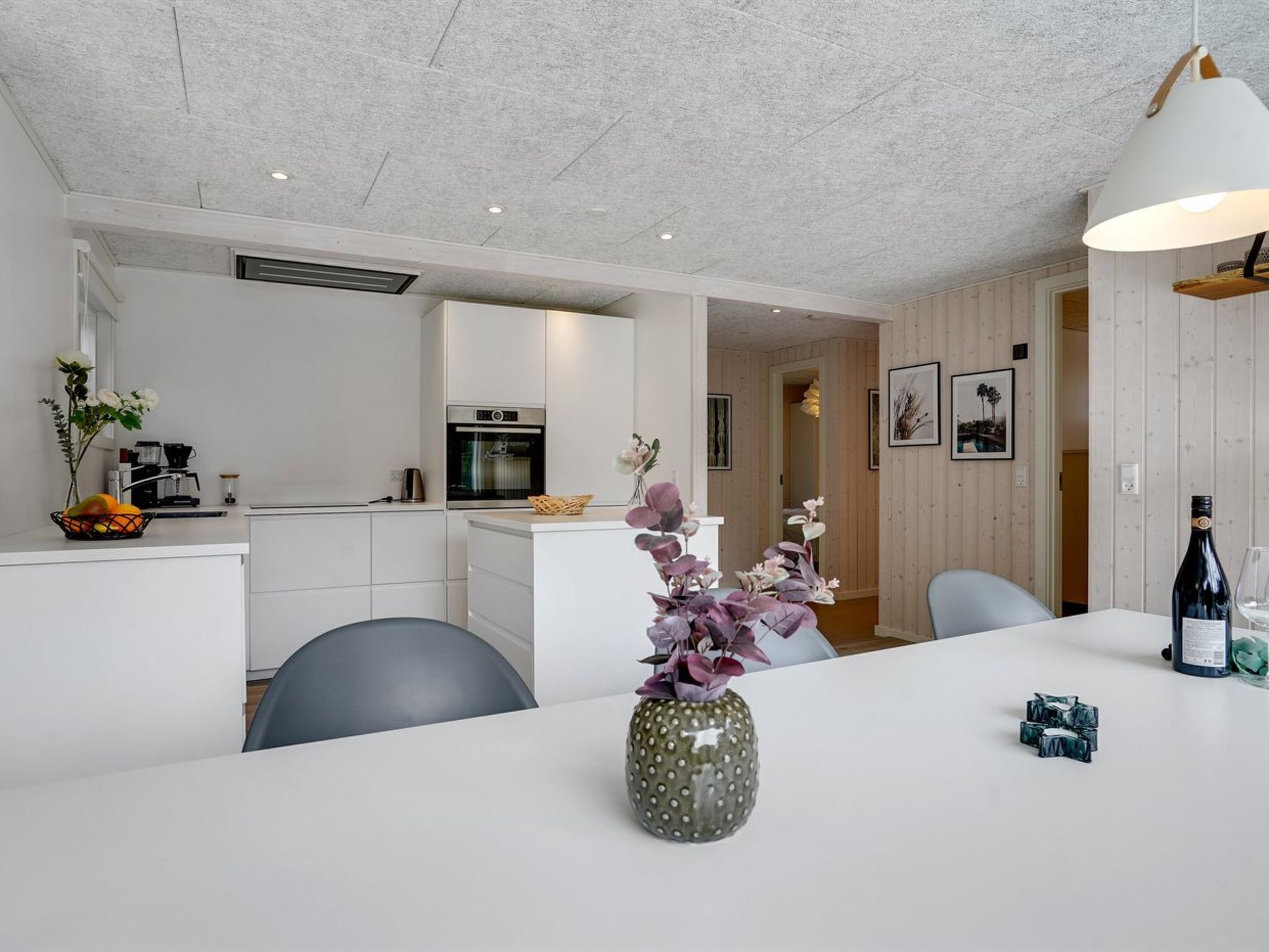 A modern kitchen with white cabinets, a built-in oven, and a coffee machine. There is a dining area with a white table, chairs, and a vase with flowers.