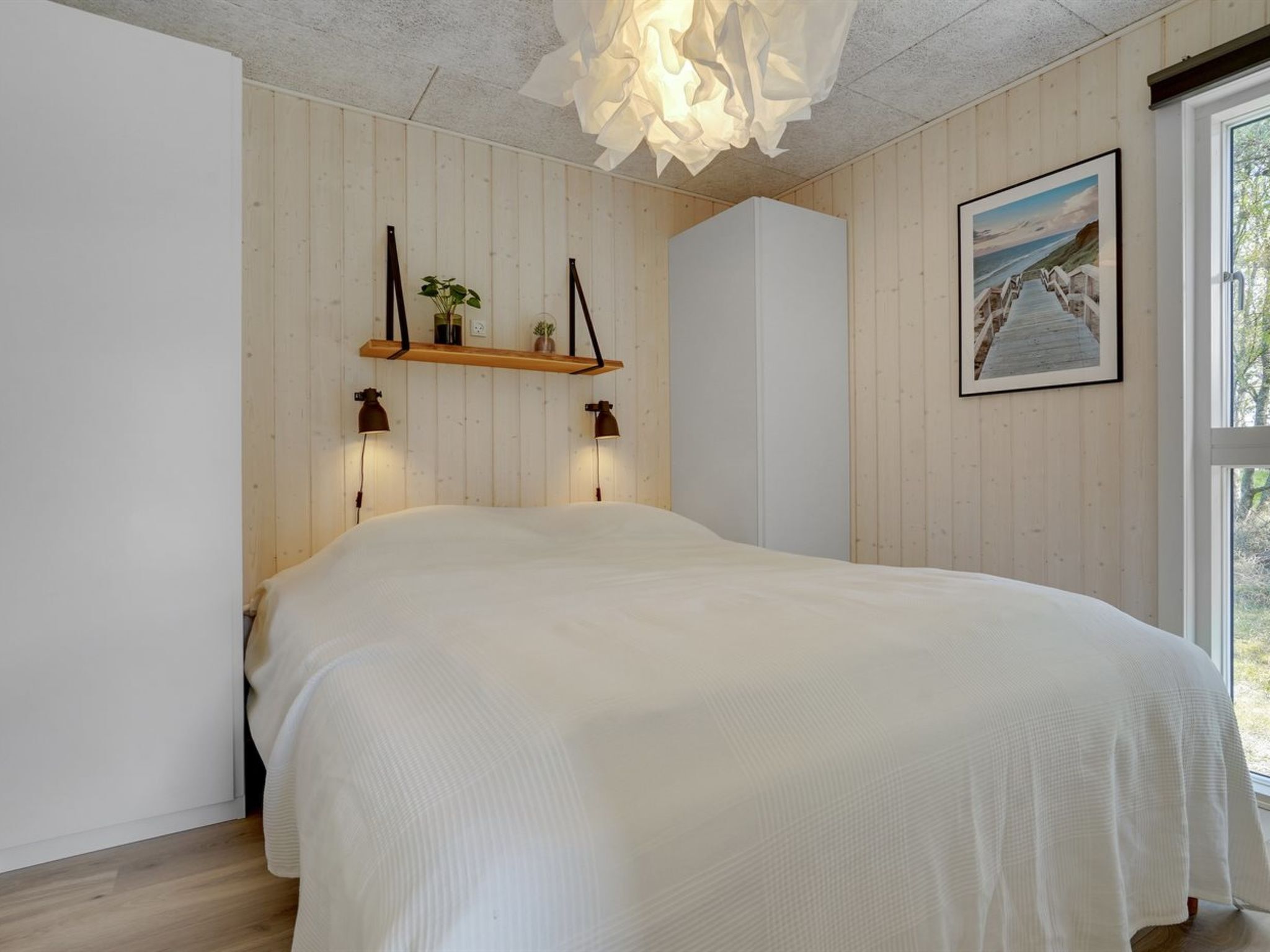 A bedroom with a large bed, wooden walls, a hanging light fixture, a shelf with plants, and a framed picture on the wall.