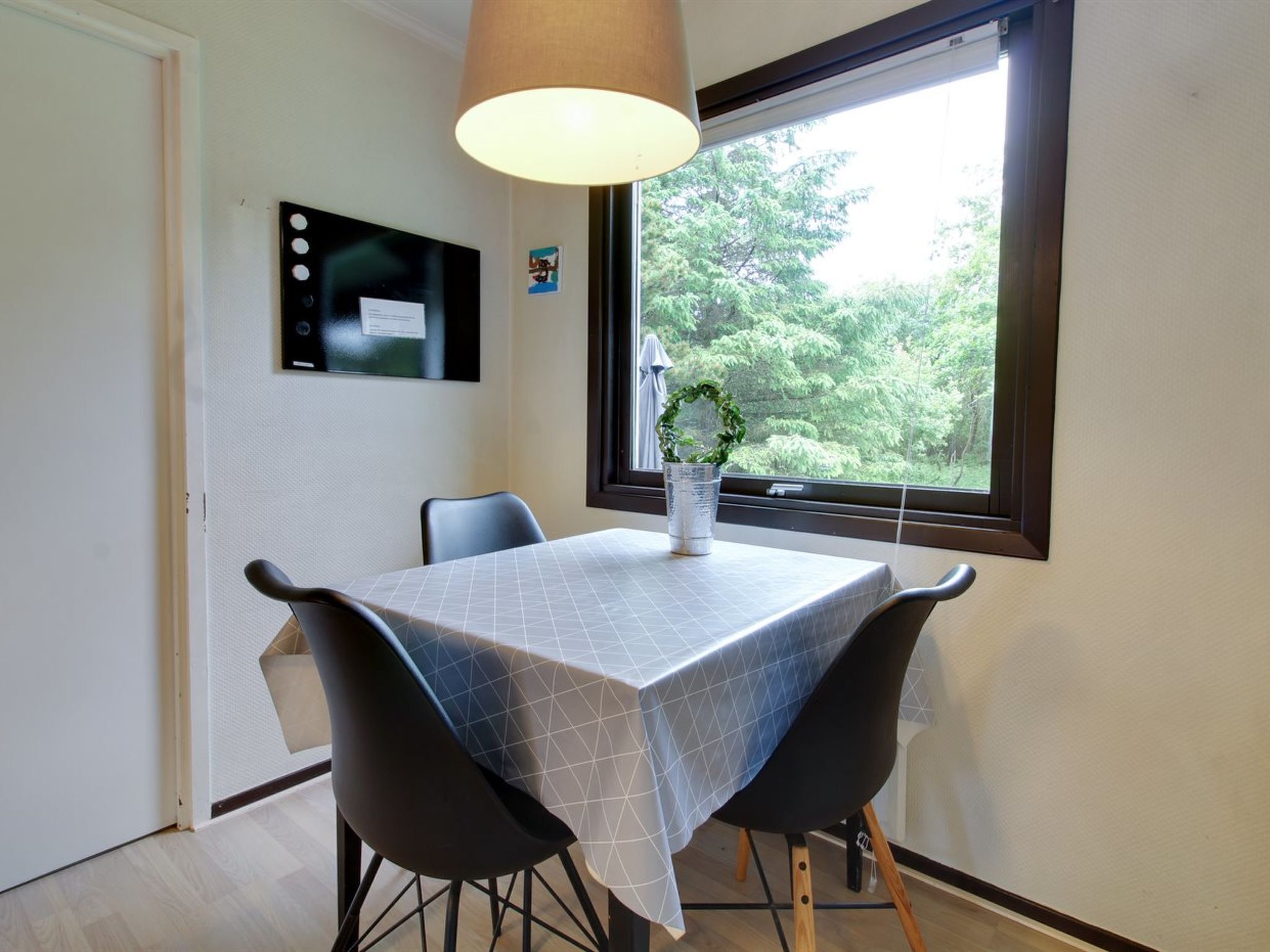 A dining area with a table covered in a geometric-patterned tablecloth, two black chairs, a hanging lamp, and a window with a view of greenery.