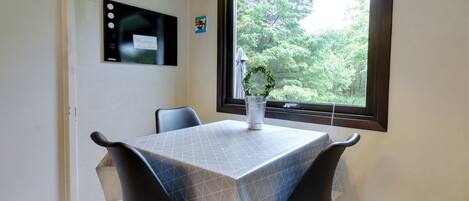 A dining area with a table covered in a geometric-patterned tablecloth, two black chairs, a hanging lamp, and a window with a view of greenery.