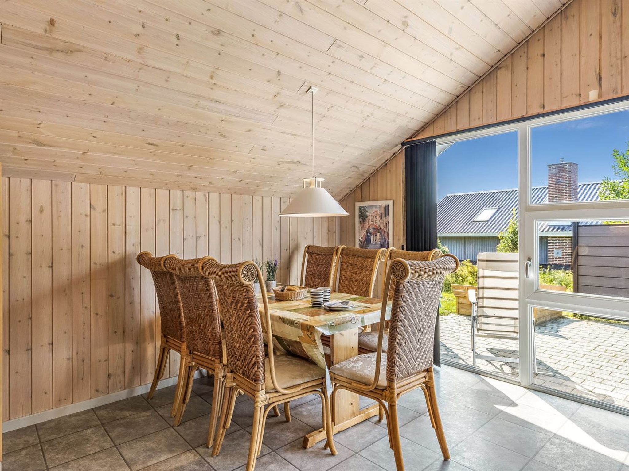 A dining area with a wooden ceiling, wicker chairs, a table with a white lamp, and a view of the outside.