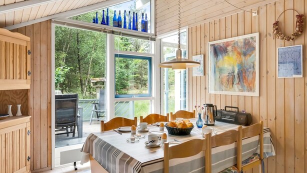 A cozy dining area with a wooden table set for four, a large window with blue glass bottles, and a painting on the wall.
