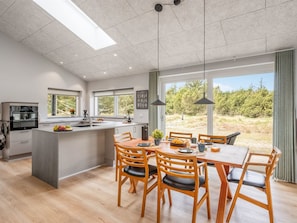 A modern kitchen with a central island, dining area with wooden chairs, large windows with a view of greenery, and pendant lighting.
