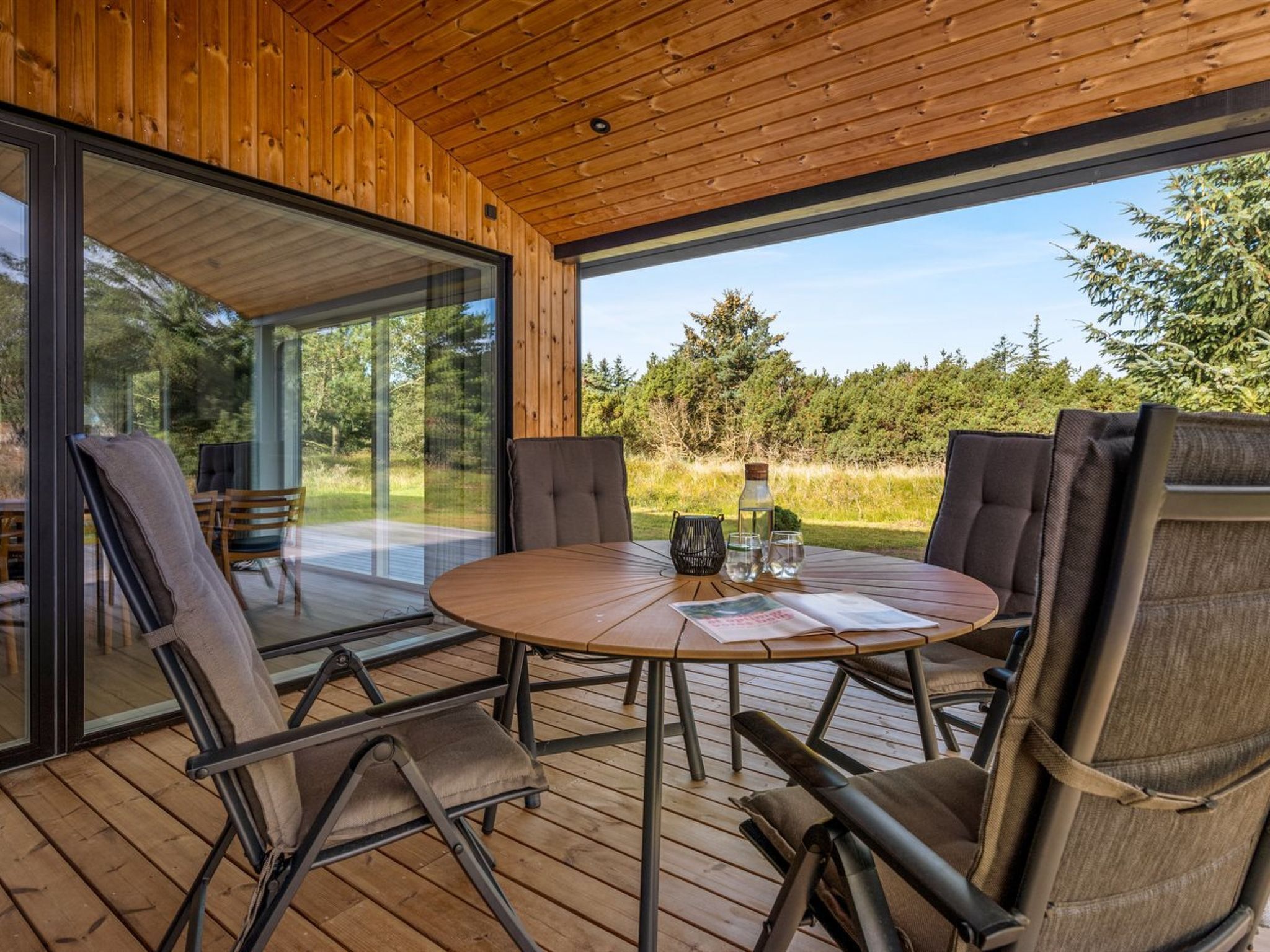 Una terraza de madera con una mesa redonda y cuatro sillas, con vista a un paisaje boscoso.