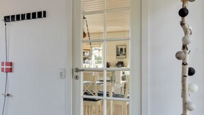 Une pièce avec une porte blanche, une petite table entourée de chaises et une étagère fixée au mur, ornée d’objets décoratifs.