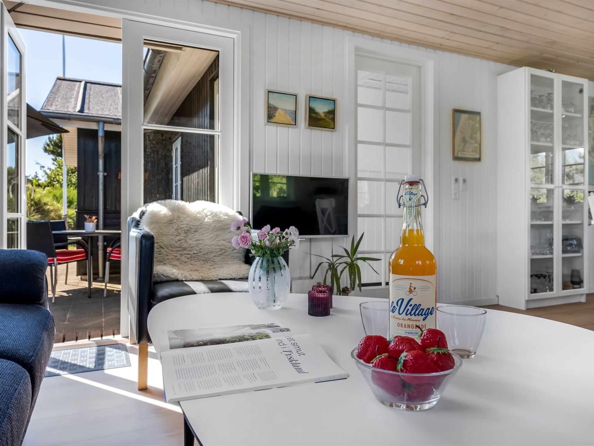 Un salon moderne avec un canapé blanc, une table basse sur laquelle se trouve une bouteille de jus d’orange, un bol de fraises et un magazine.