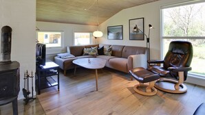 Ein modernes Wohnzimmer mit Holzbalkendecke, einer Couch, einem Couchtisch, einem Kamin und einem großen Fenster mit Blick auf Bäume.