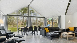 Un vaste espace intérieur doté d’un haut plafond, de grandes fenêtres, d’un canapé en cuir noir, d’une table à manger avec des chaises, et d’un espace de jeux visible à travers les fenêtres.