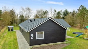 Une petite cabane de couleur sombre, portant le numéro « 2 » sur le côté, entourée d’une cour verdoyante et d’une aire de jeux.