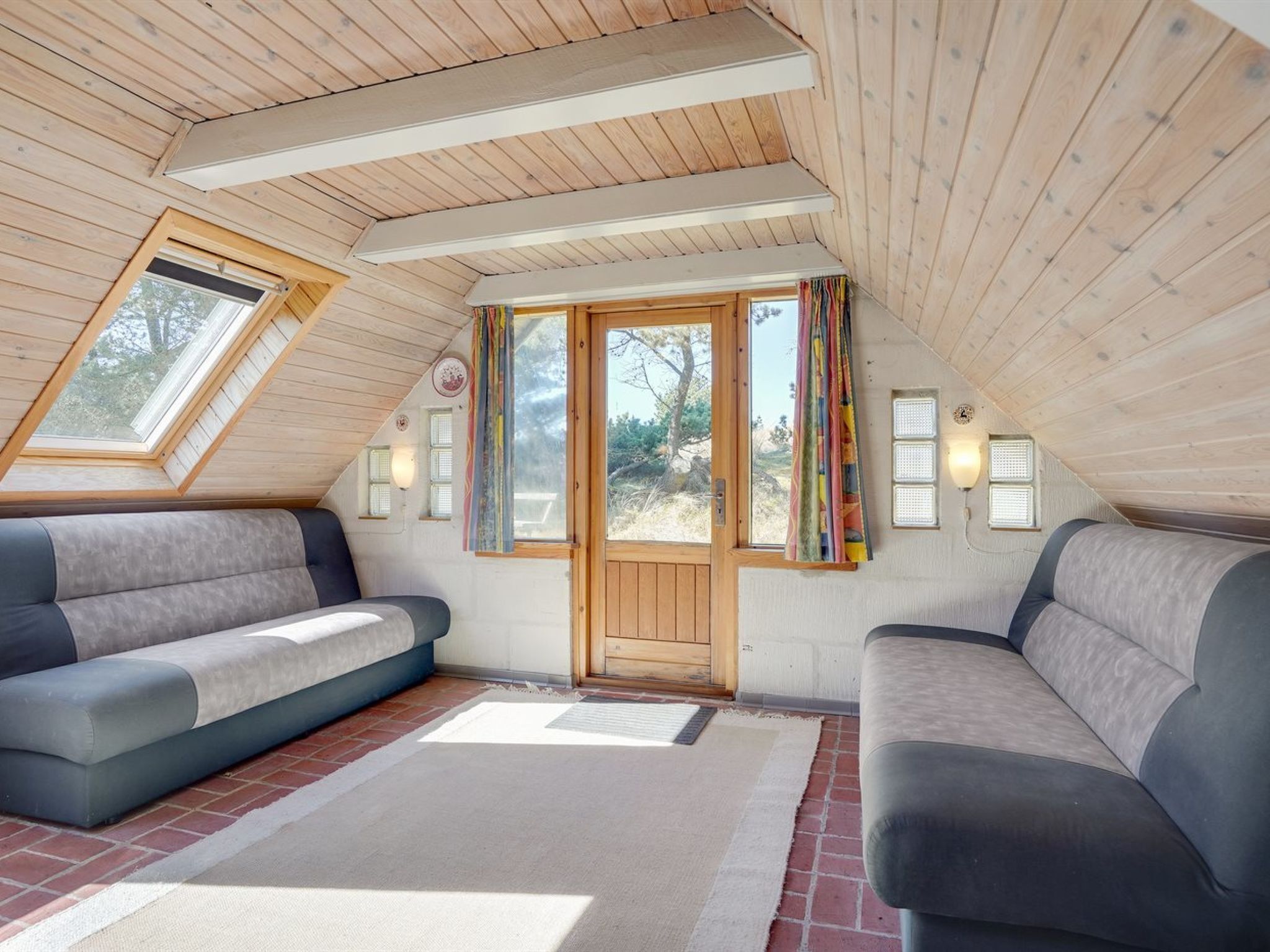 Une chambre mansardée chaleureuse, avec deux canapés, un plafond en bois et une grande fenêtre agrémentée de rideaux.