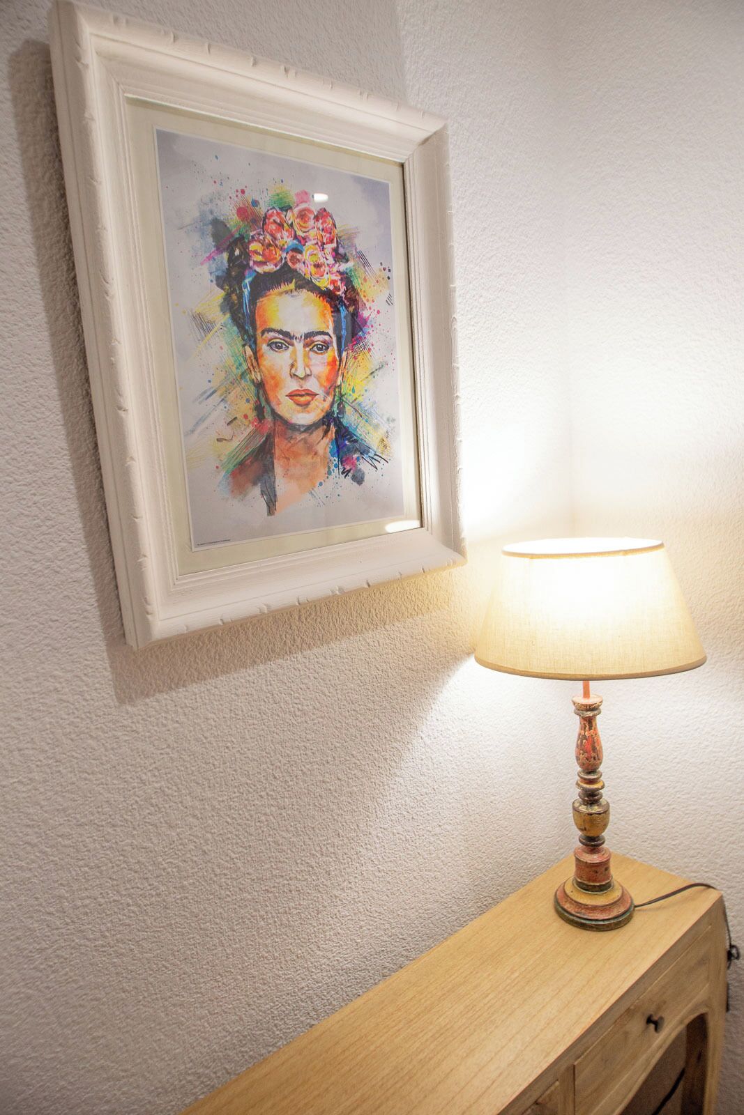 Een lijstwerk aan de muur, een lamp op een houten bijzettafeltje, en een vloer met tapijt.