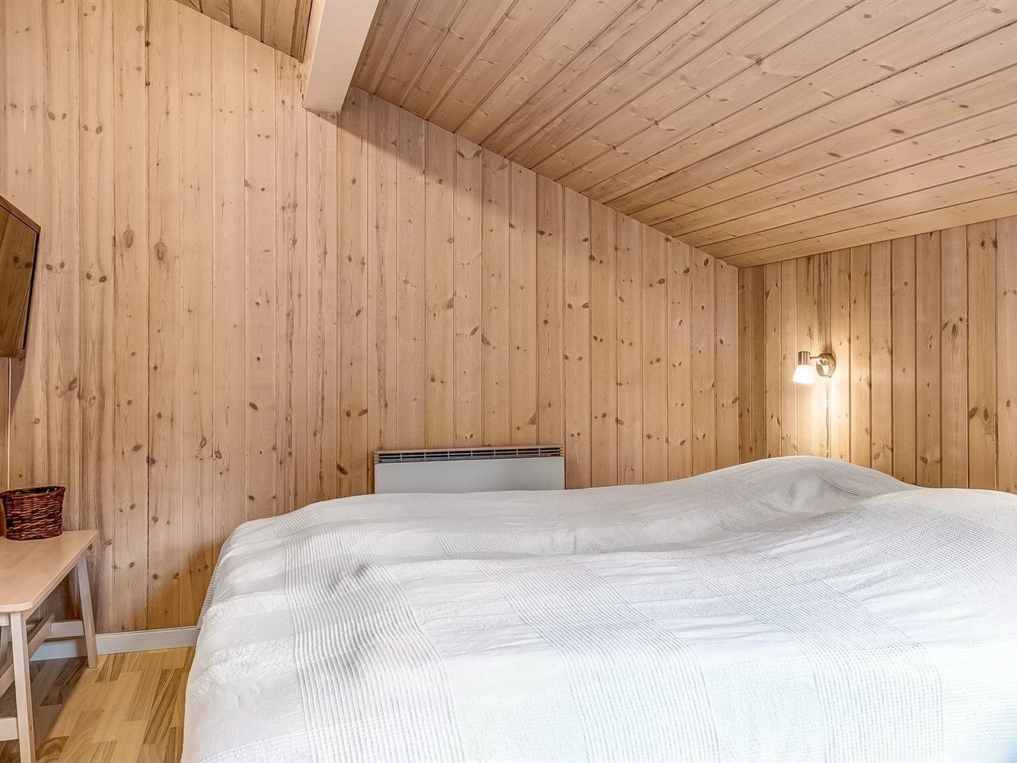 Et soveværelse med trævægge og -loft, en seng med hvid dyne, et natbord med en kurv og en vægmonteret lampe.