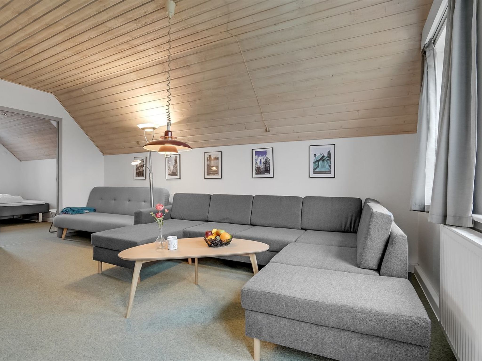 Et moderne loftværelse med skråvægge, en grå hjørnesofa, et lille rundt bord med en vase blomster og en gulvlampe.