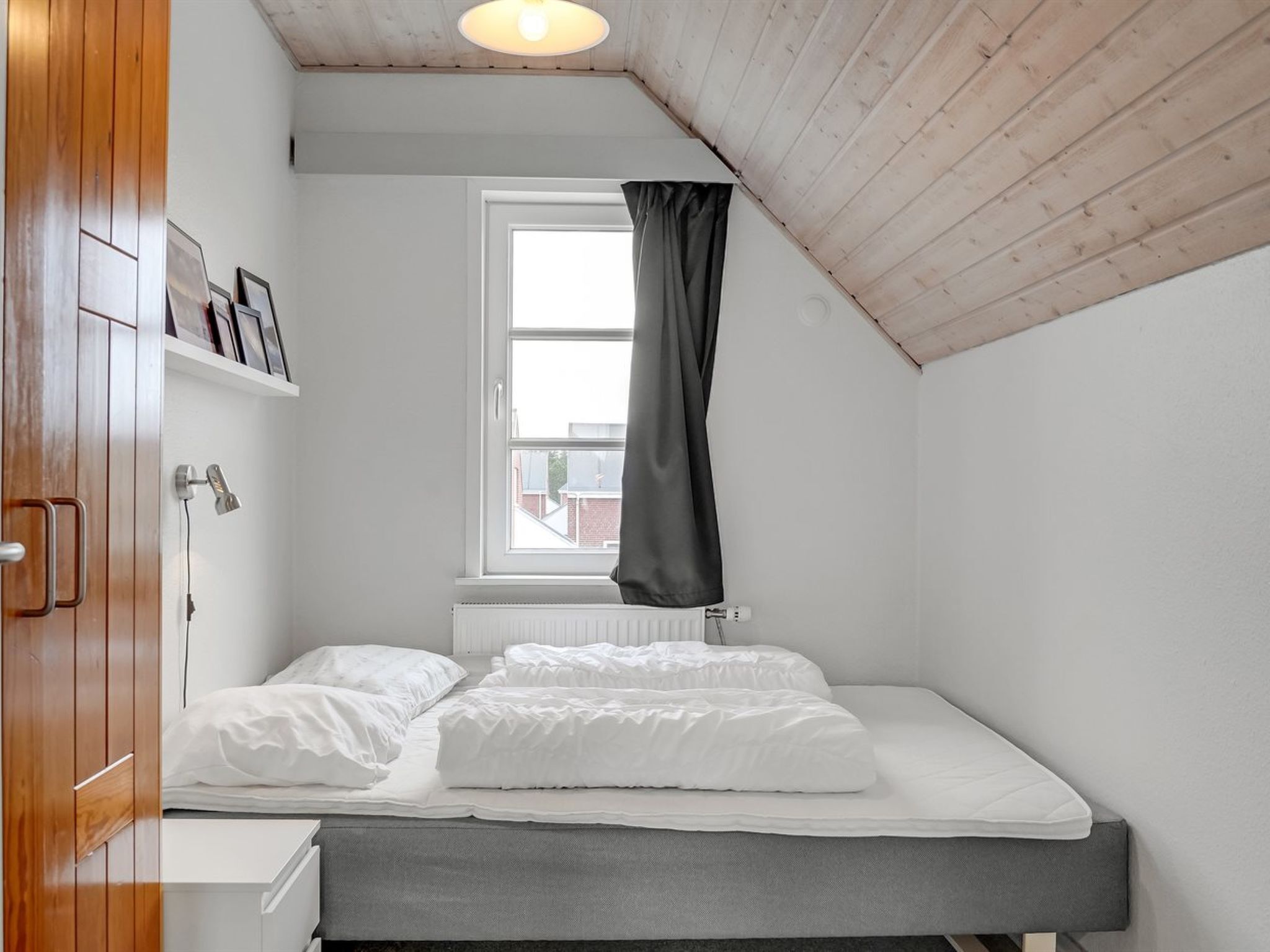A small, single-bed room with a sloped ceiling, a window with a black curtain, a white radiator, and a wooden wardrobe with a shelf above it.