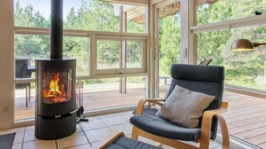 Une pièce chaleureuse avec un poêle à bois, un fauteuil confortable et de grandes fenêtres donnant sur le paysage extérieur.